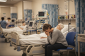A middle-aged woman sitting beside her mother in a hospital emergency room, face in her hands, overwhelmed and trying to figure out what to do next.